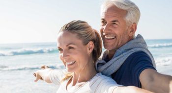 Happy senior couple standing on beach with arms outstretched and looking away. Happy couple at beach on a bright sunny day. Retired husband and smiling wife thinking about their future.