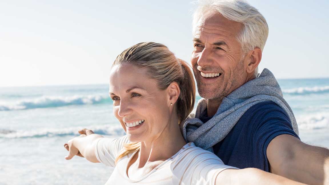 Happy senior couple standing on beach with arms outstretched and looking away. Happy couple at beach on a bright sunny day. Retired husband and smiling wife thinking about their future.