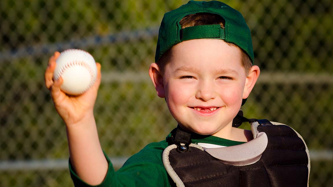 Young child in catcher's gear throwing baseball while laughing