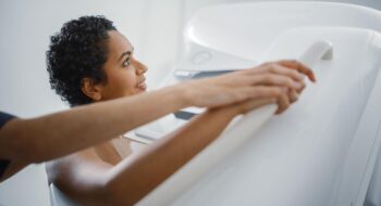 Friendly Female Doctor Explains the Mammogram Procedure to a Topless Latin Female Patient with Curly Hair Undergoing Mammography Scan. Healthy Female Does Cancer Prevention Routine in Hospital Room.