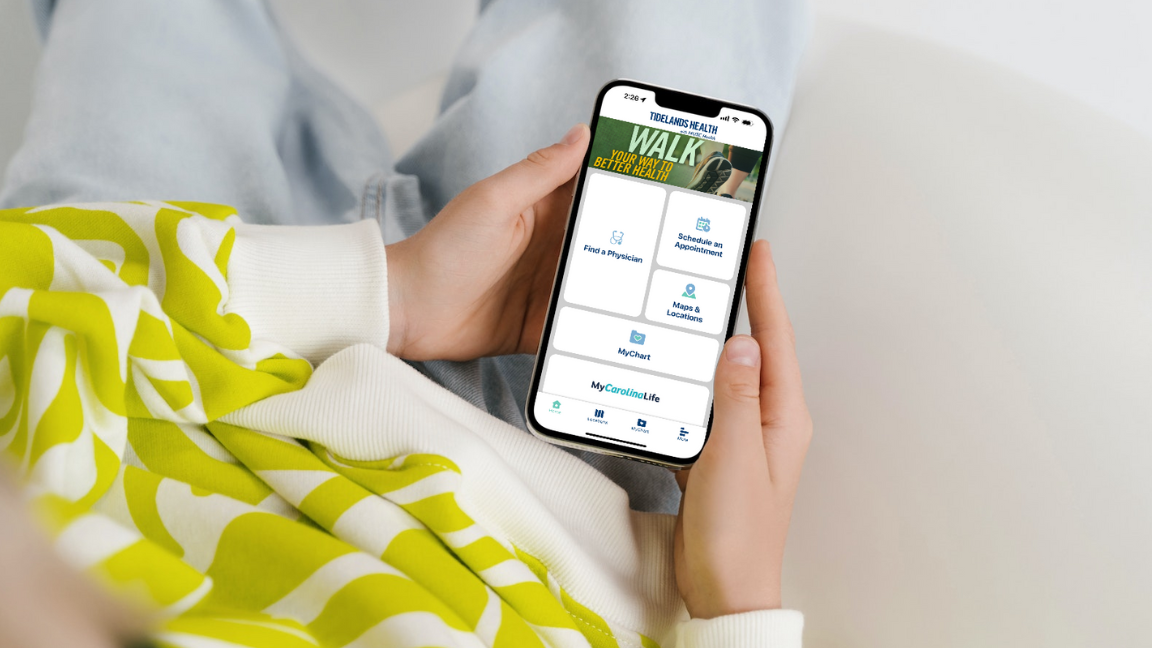 A person holding a smartphone in their lap. The phone shows My Tidelands Health app on the screen.