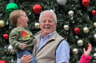 An older man with a child in front of a Christmas tree. Everyone is smiling.