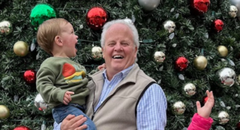 An older man with a child in front of a Christmas tree. Everyone is smiling.