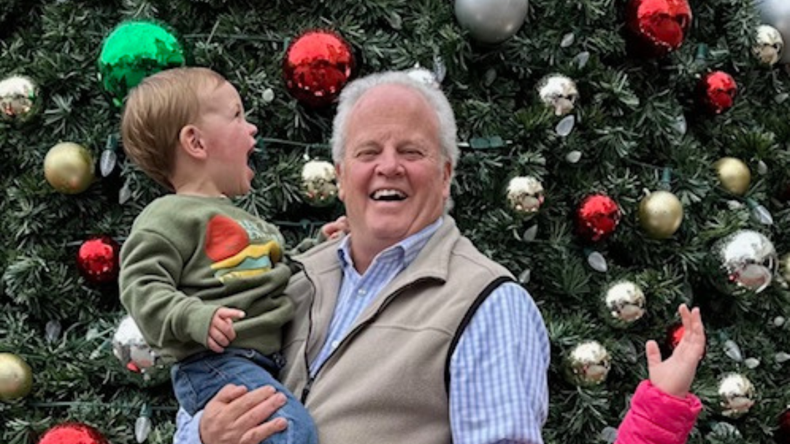 An older man with a child in front of a Christmas tree. Everyone is smiling.