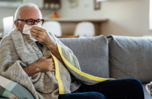 A senior Caucasian man is lying in bed with a bad cold and is blowing his nose using a napkin.