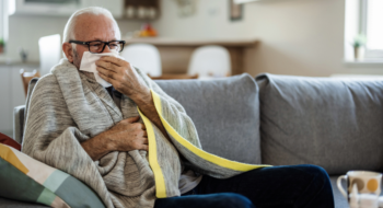 A senior Caucasian man is lying in bed with a bad cold and is blowing his nose using a napkin.