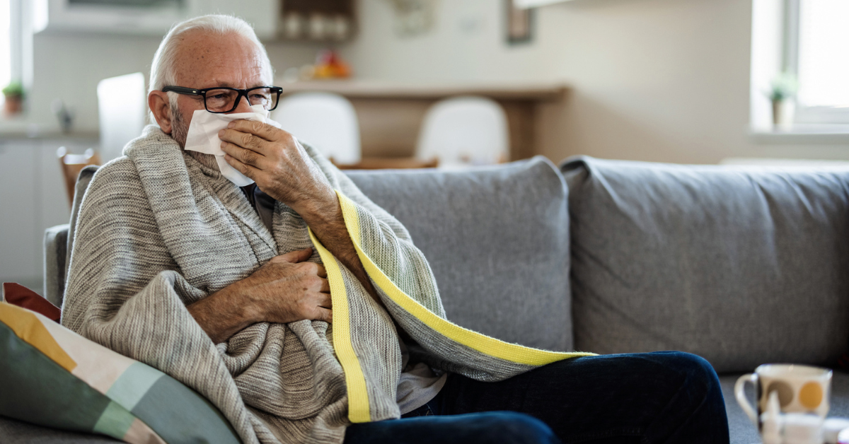 A senior Caucasian man is lying in bed with a bad cold and is blowing his nose using a napkin.