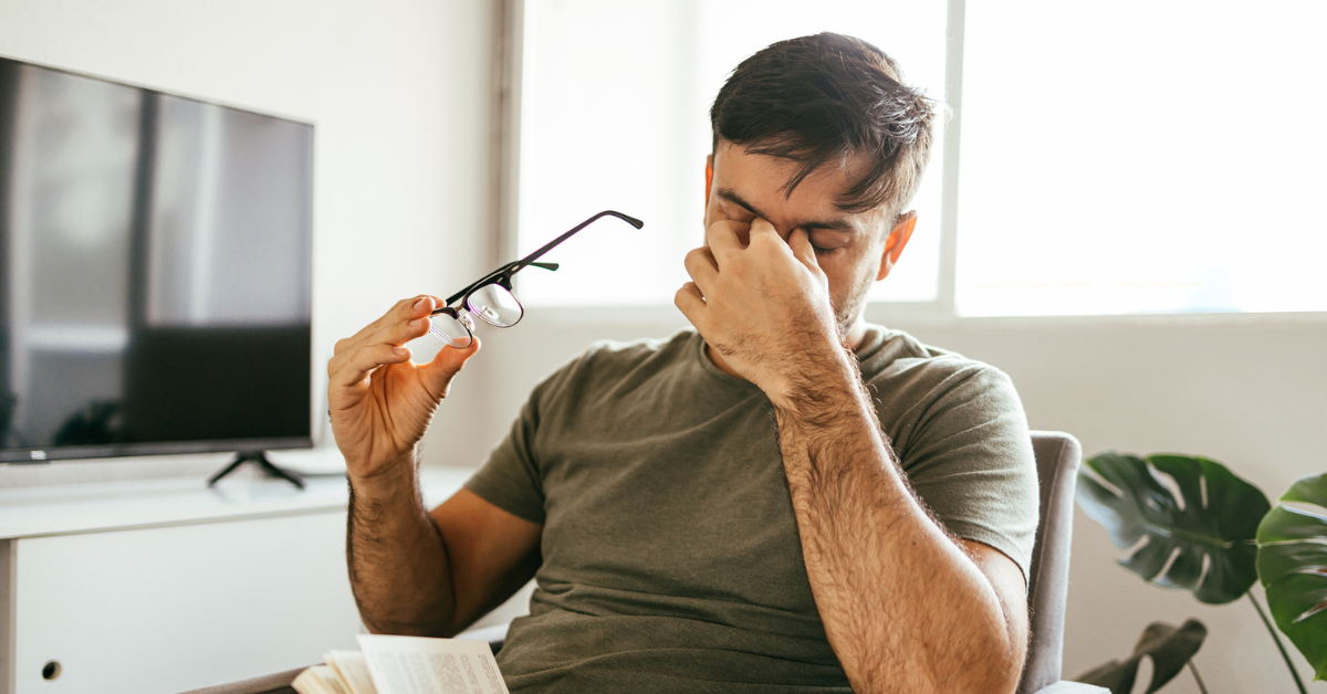 Tired man takes off his glasses and rubs his eyes while reading a book at home.