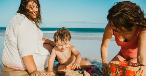 Adults smiling and watching cute boy building castle from wet sand while resting on beach near sea together