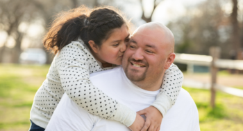 Daughter hugging her dad from behind and kissing his cheek.