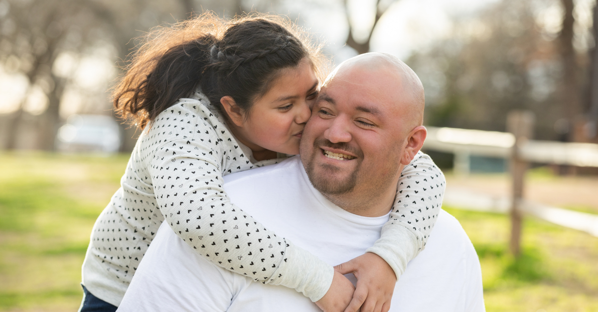 Daughter hugging her dad from behind and kissing his cheek.