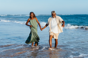 A cheerful older couple holds hands while walking and laughing together along the sandy beach, embodying joy and togetherness.
