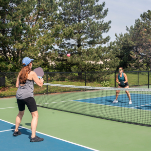 A comptetivie doubles game of pickleball at the net on a blue and green court in summer.