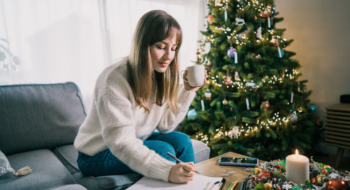 A woman sits on her couch writing down new year resolutions while holding a coffee mug in her hand.