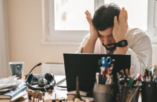 A man sitting at his desk with his computer rubbing his head in frustration.