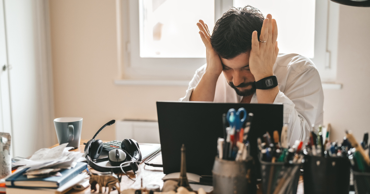 A man sitting at his desk with his computer rubbing his head in frustration.