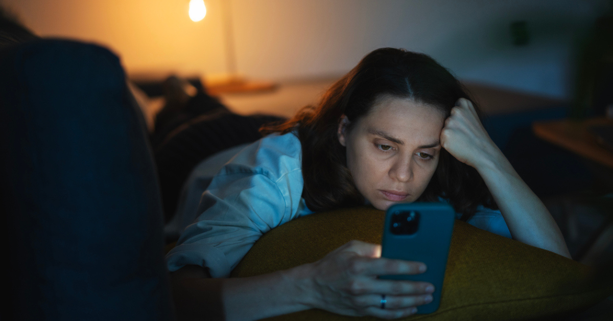 Young serious sad woman lying on sofa and looking at smartphone screen at night or evening