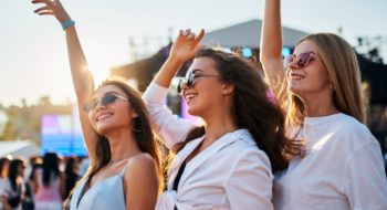 Arms raised, dancing in evening light. Group of three young women enjoying summer music festival on sandy shore. Fashionable friends party, carefree atmosphere with ocean backdrop on warm day.