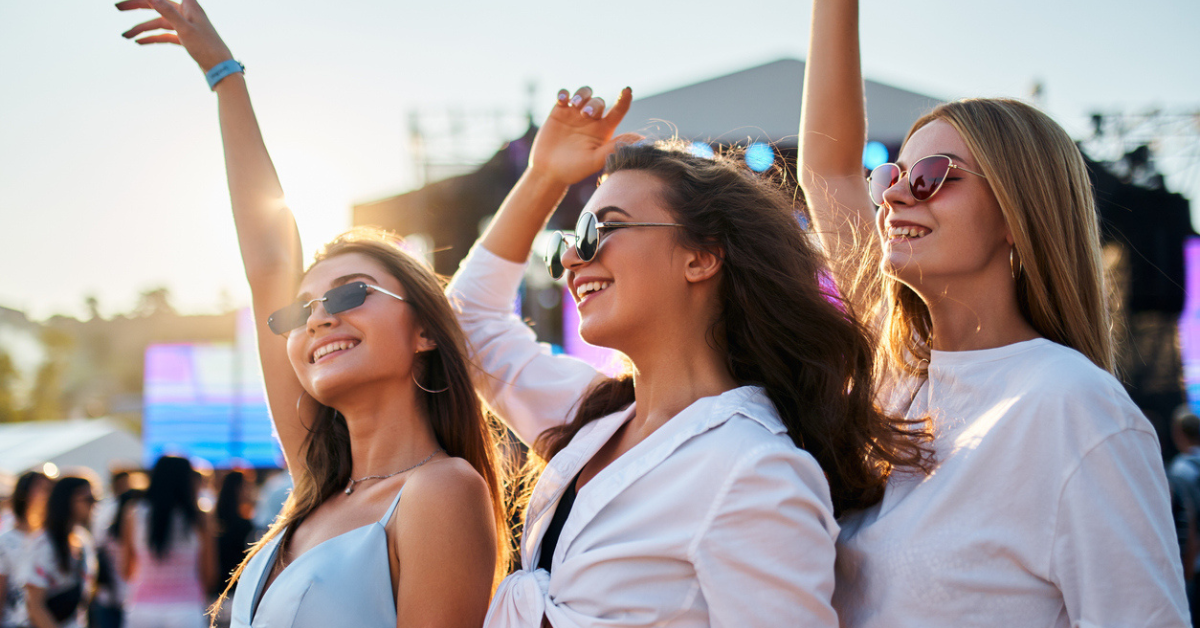 Arms raised, dancing in evening light. Group of three young women enjoying summer music festival on sandy shore. Fashionable friends party, carefree atmosphere with ocean backdrop on warm day.