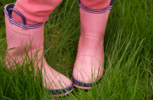 A child standing with their feet turn inward in a field of grass, wearing pink rainboots and pants.