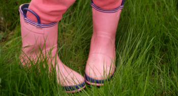 A child standing with their feet turn inward in a field of grass, wearing pink rainboots and pants.