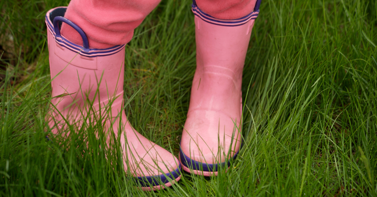 A child standing with their feet turn inward in a field of grass, wearing pink rainboots and pants.