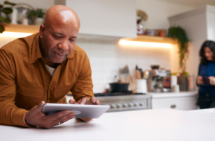 Mature Man At Home In Kitchen Looking At Digital Tablet With Female Partner In Background