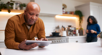 Mature Man At Home In Kitchen Looking At Digital Tablet With Female Partner In Background