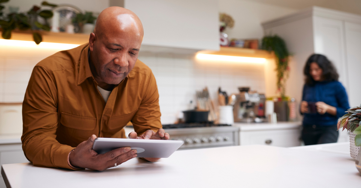 Mature Man At Home In Kitchen Looking At Digital Tablet With Female Partner In Background