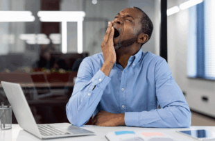 Mature African American businessman yawning at office desk with open laptop. Depicts workplace fatigue, stress, and need for rest. Blue shirt contrasts modern professional environment.