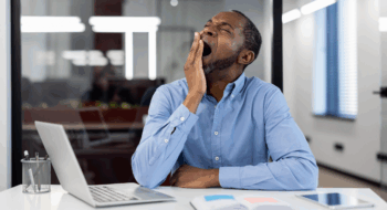 Mature African American businessman yawning at office desk with open laptop. Depicts workplace fatigue, stress, and need for rest. Blue shirt contrasts modern professional environment.