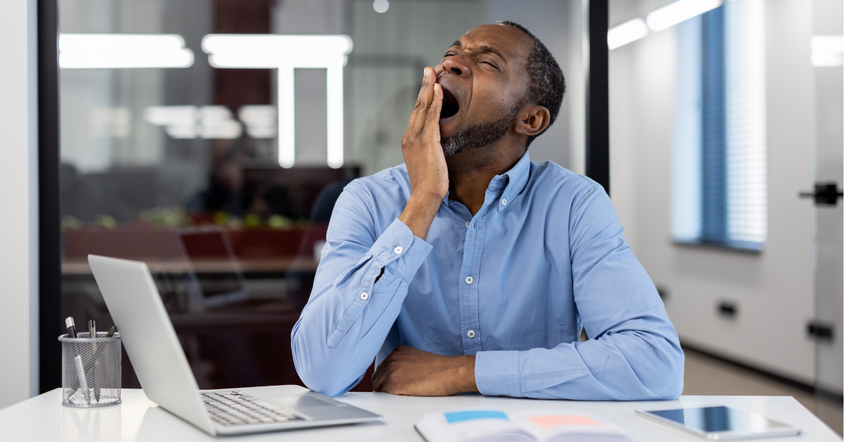 Mature African American businessman yawning at office desk with open laptop. Depicts workplace fatigue, stress, and need for rest. Blue shirt contrasts modern professional environment.