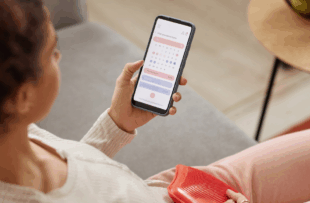 High angle closeup of young woman holding smartphone with period tracker app on screen and holding hot water bottle on belly