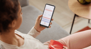 High angle closeup of young woman holding smartphone with period tracker app on screen and holding hot water bottle on belly