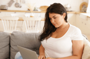 plus size student girl with loose black hair keyboarding on laptop sitting comfortably on sofa in living room