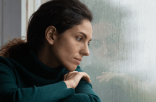 Thoughtful stressed young hispanic latin woman sitting on windowsill, looking outside on rainy weather, having depressive or melancholic mood, suffering from negative thoughts alone at home.