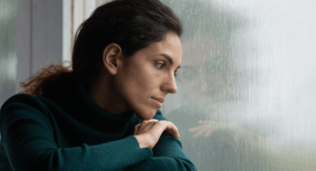 Thoughtful stressed young hispanic latin woman sitting on windowsill, looking outside on rainy weather, having depressive or melancholic mood, suffering from negative thoughts alone at home.