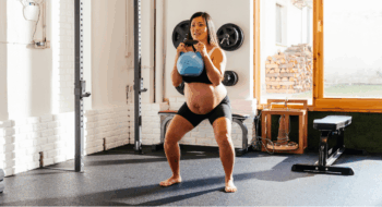 A pregnant woman is engaged in a kettlebell squat in a well-lit home gym.