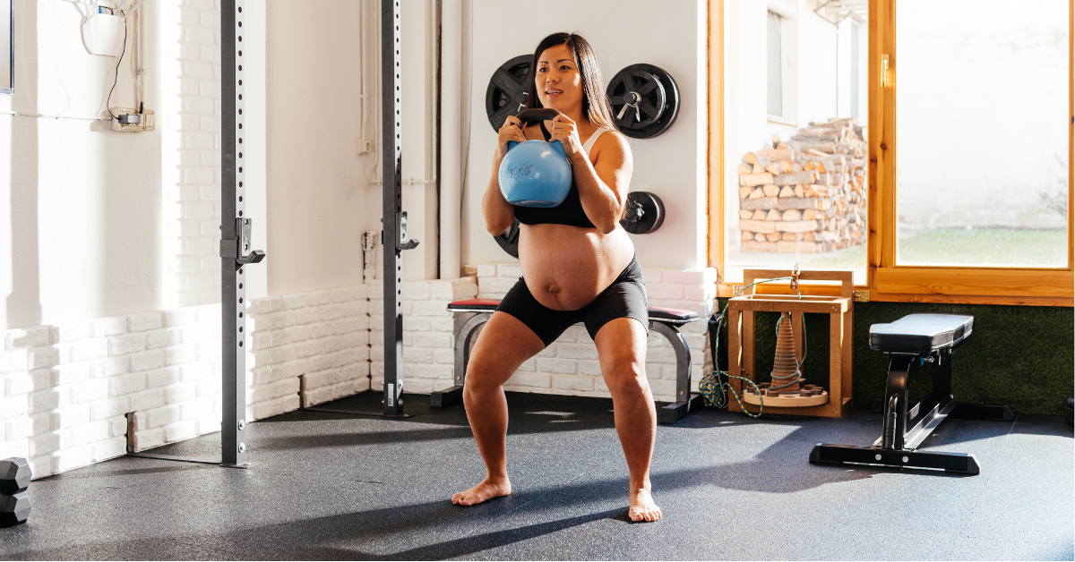 A pregnant woman is engaged in a kettlebell squat in a well-lit home gym.
