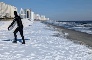 A surfer coming out of the ocean wearing a black wetsuit and holding a white surfboard. There is snow on the beach.