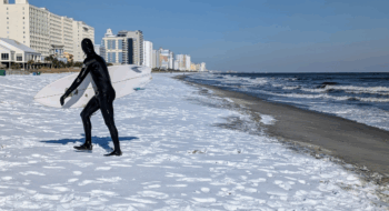 A surfer coming out of the ocean wearing a black wetsuit and holding a white surfboard. There is snow on the beach.
