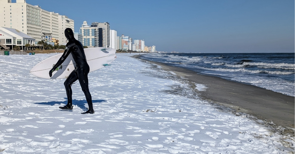 A surfer coming out of the ocean wearing a black wetsuit and holding a white surfboard. There is snow on the beach.