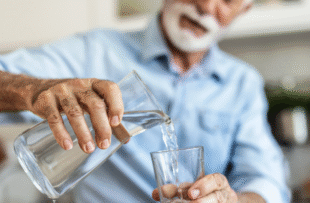 An elderly man in a blue button down shirt pouring water into a glass from a glass pitcher.