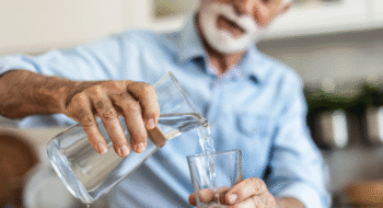 An elderly man in a blue button down shirt pouring water into a glass from a glass pitcher.