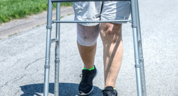A real person, real life recent knee replacement surgery patient senior adult woman is doing her prescribed physical therapy. For safety and support, she uses an orthopedic mobility walker to walk slowly and carefully for increasing daily distances on the quiet streets around her suburban home. The open mesh bandage around her right knee protects and supports the surgical site wound and stitches while allowing constant fresh air to help the healing process.
