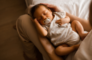 Sleeping baby boy 1-2 months old with pacifier sleeping on mother hands in bedroom closeup top view.