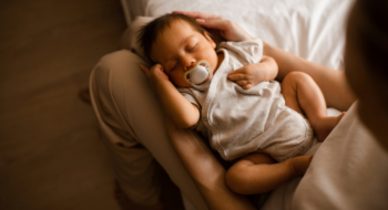Sleeping baby boy 1-2 months old with pacifier sleeping on mother hands in bedroom closeup top view.
