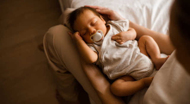 Sleeping baby boy 1-2 months old with pacifier sleeping on mother hands in bedroom closeup top view.