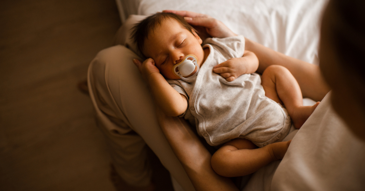 Sleeping baby boy 1-2 months old with pacifier sleeping on mother hands in bedroom closeup top view.